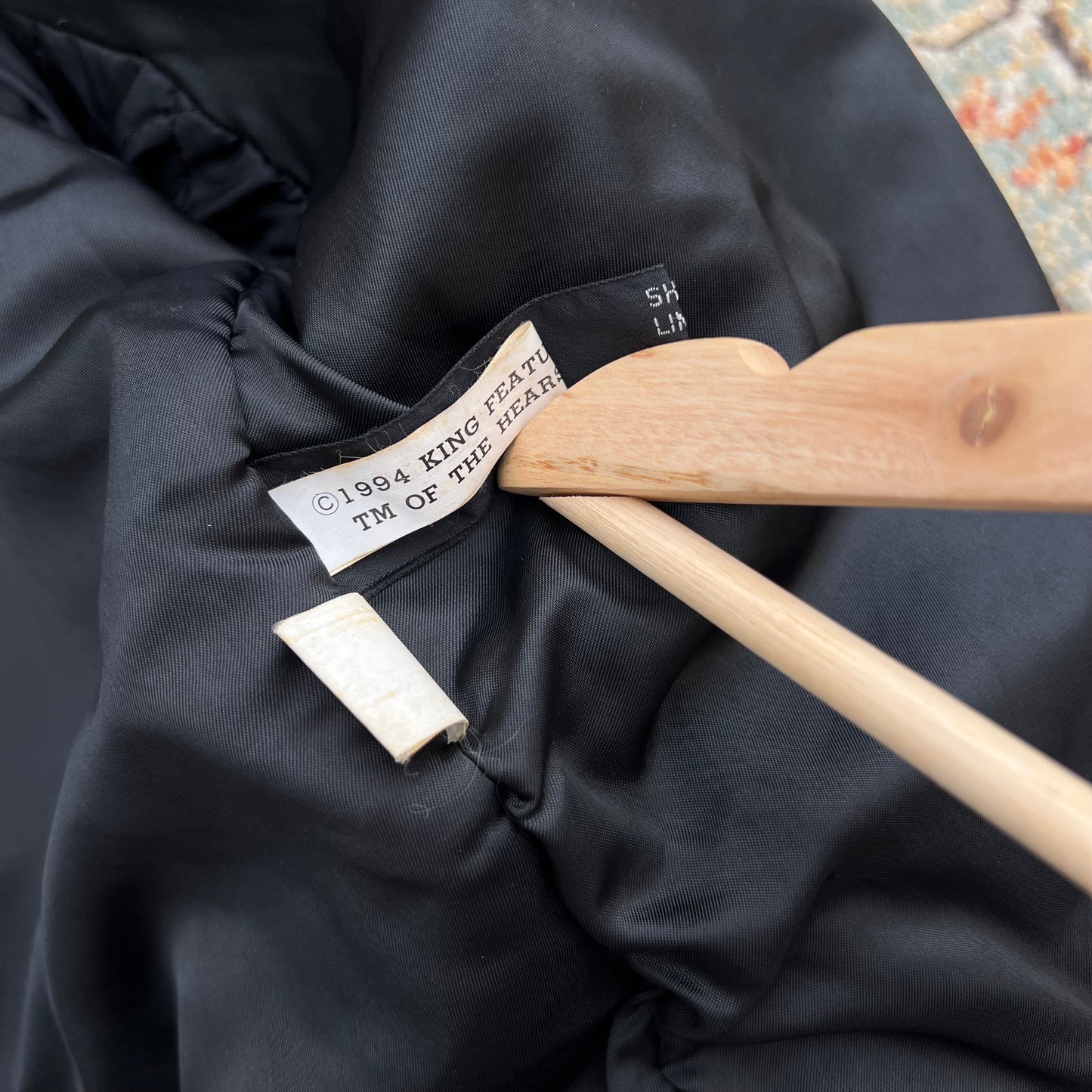 Close-up of a black puffer jacket interior label reading '1994 KING FEAT TM OF THE SEAP' on a wooden hanger, with a small white tag nearby.