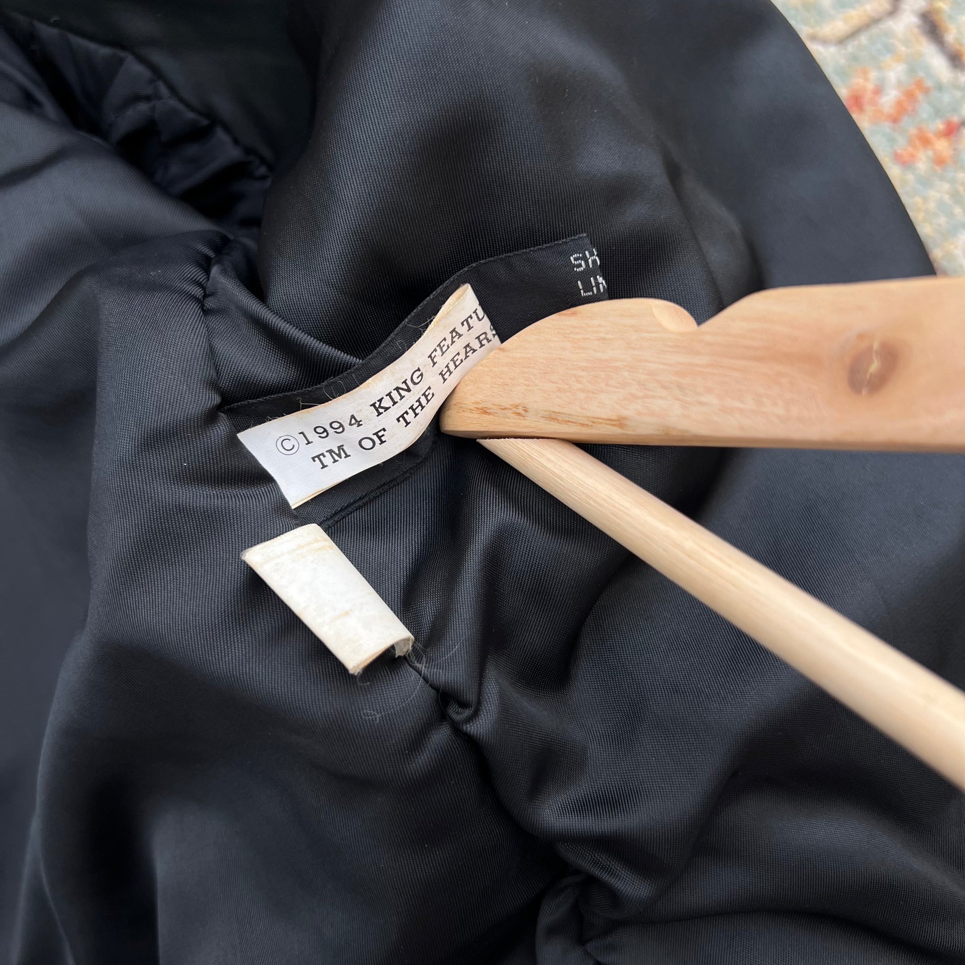 Close-up of a black puffer jacket interior label reading '1994 KING FEAT TM OF THE SEAP' on a wooden hanger, with a small white tag nearby.