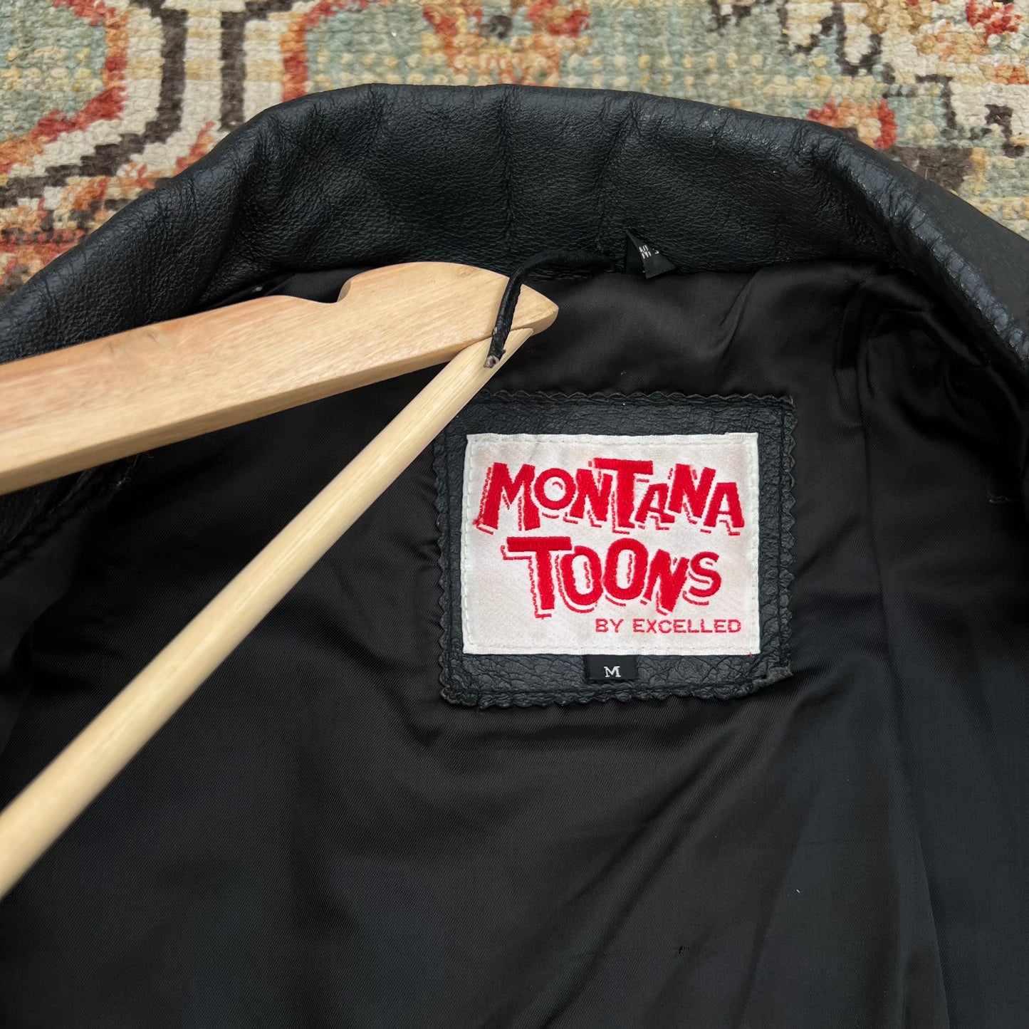 Close-up of the inside neck area of a black jacket on a wooden hanger, showing a white brand label that reads 'MONTANA TOONS BY EXCELLED' in red lettering.