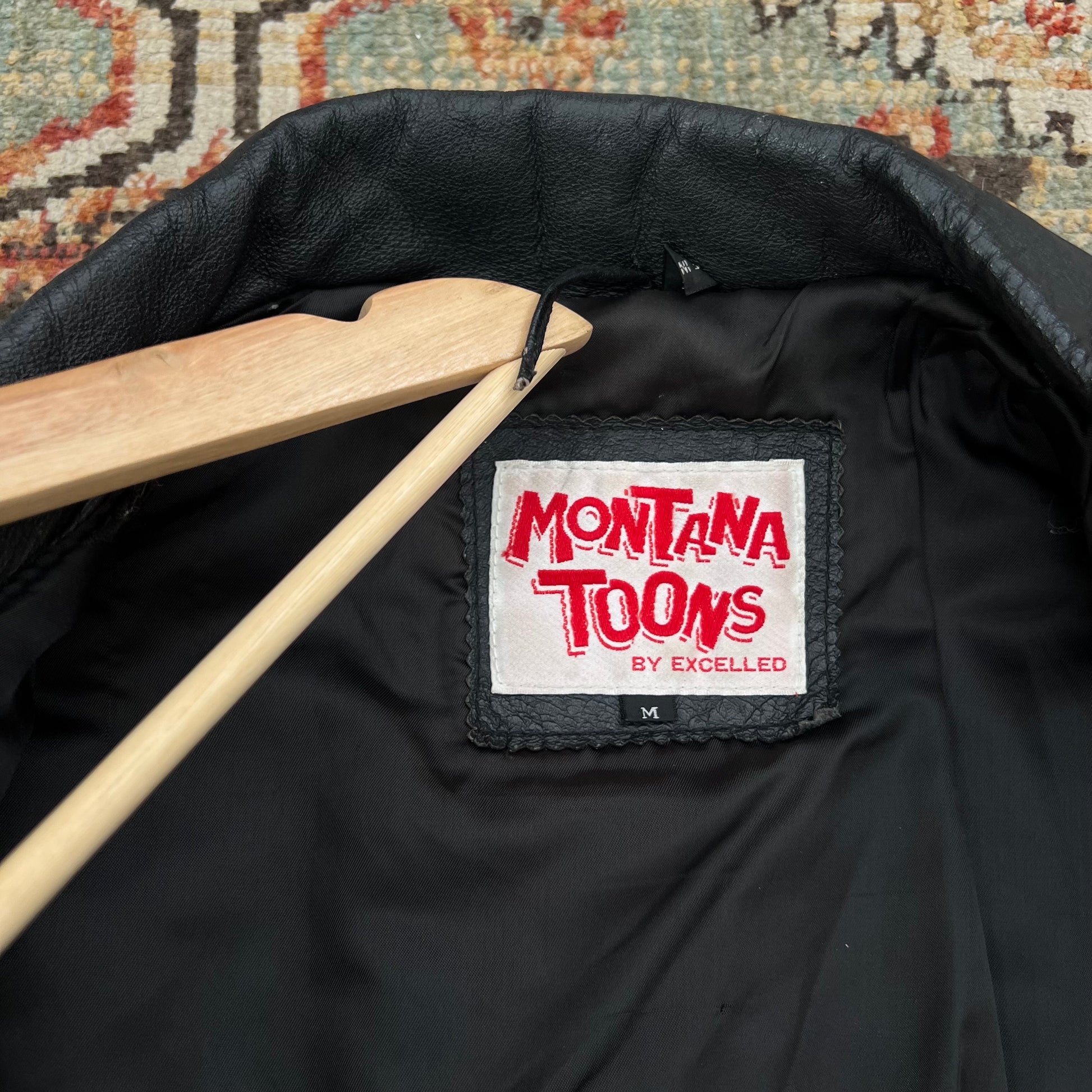 Close-up of the inside neck area of a black jacket on a wooden hanger, showing a white brand label that reads 'MONTANA TOONS BY EXCELLED' in red lettering.
