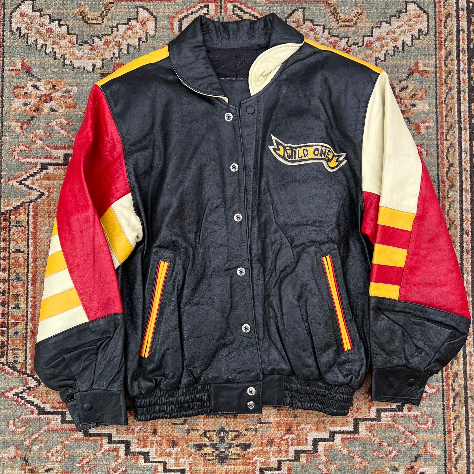 A vintage black leather varsity jacket with red and cream color-blocked sleeves, featuring a 'MILD ONE' logo patch on the left chest, laid flat on a patterned rug.
