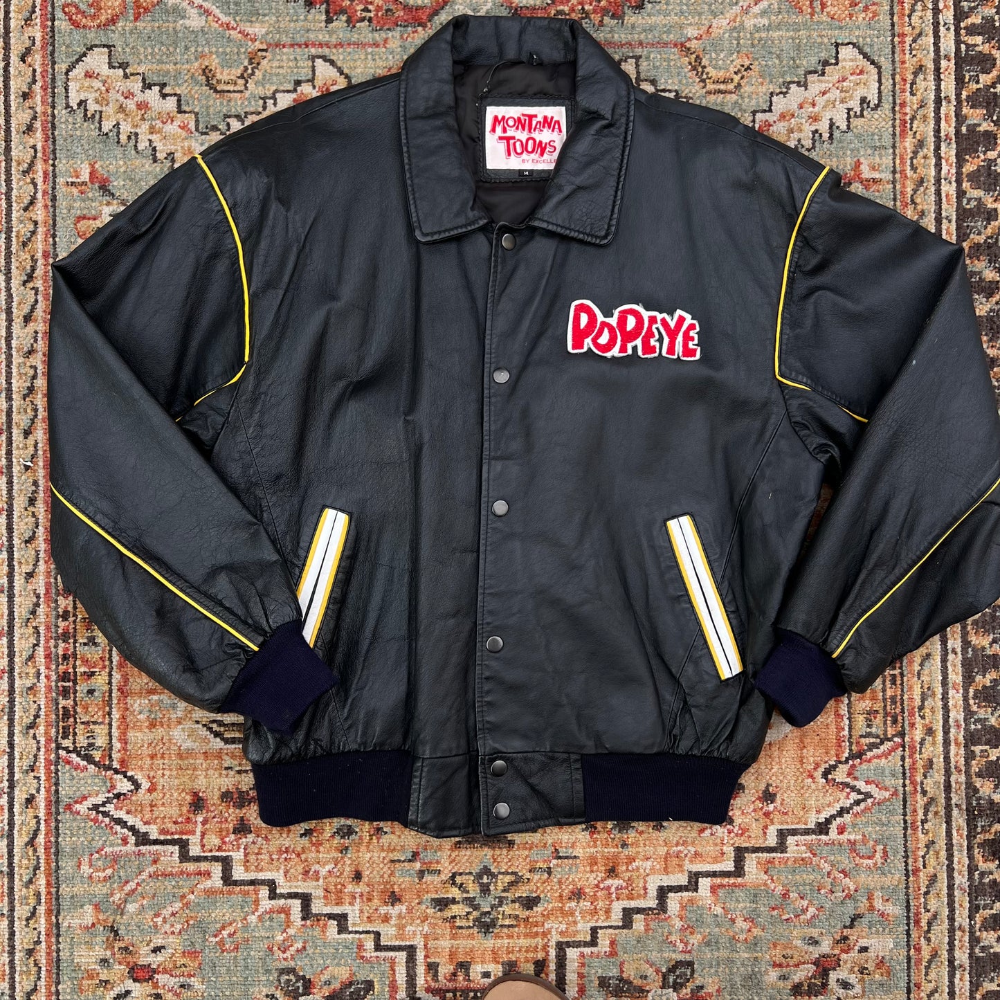 A black leather jacket with yellow piping and a red 'Popeye' logo on the chest, laid flat on a patterned rug. The jacket features a button-up front, ribbed collar, cuffs, and hem, and reflective yellow-and-white stripes on the pockets.