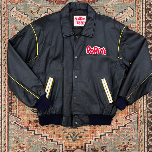 A black leather jacket with yellow piping and a red 'Popeye' logo on the chest, laid flat on a patterned rug. The jacket features a button-up front, ribbed collar, cuffs, and hem, and reflective yellow-and-white stripes on the pockets.