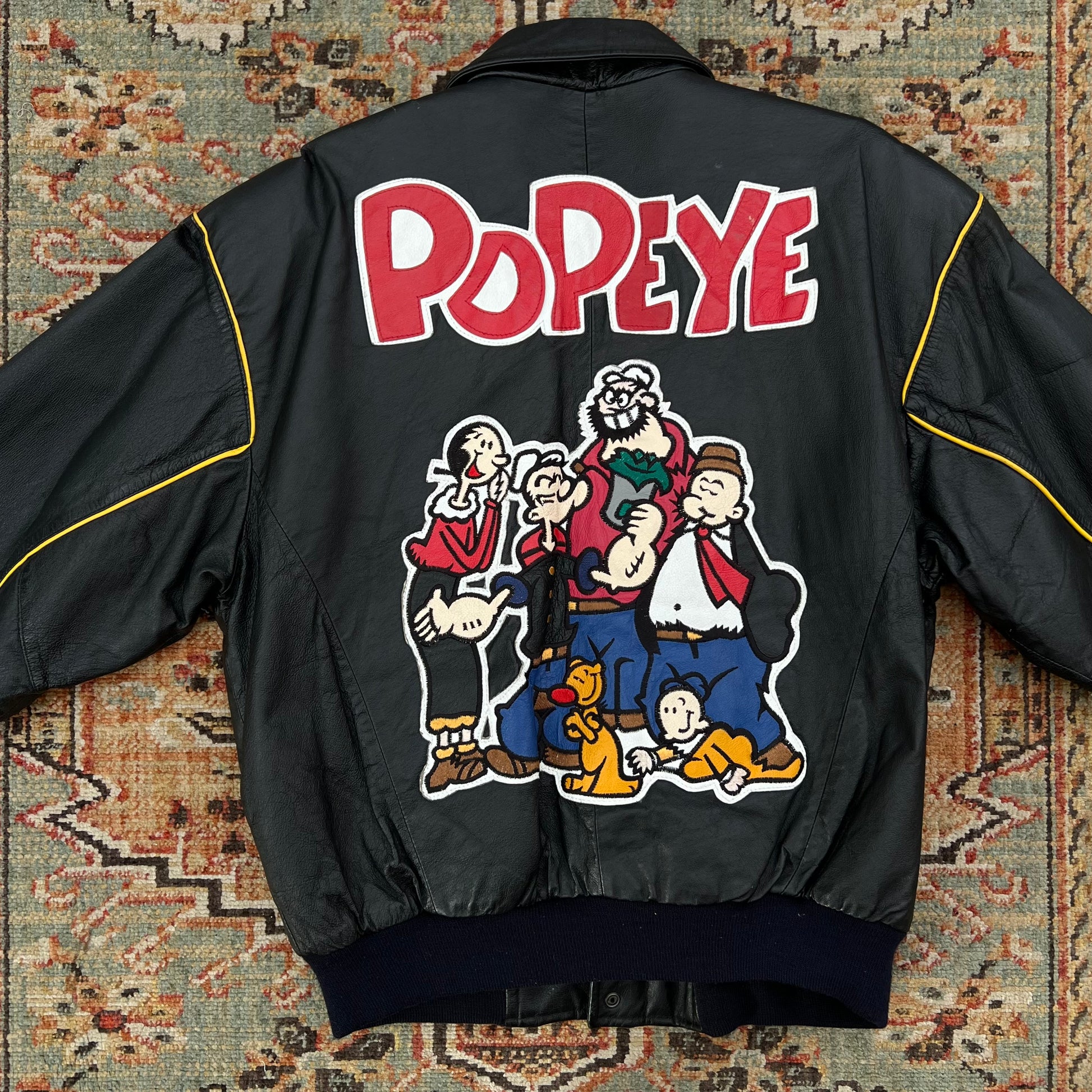 A black leather bomber jacket featuring a large back graphic of the Popeye cartoon family with the word 'Popeye' in red letters, laid flat on a patterned rug.