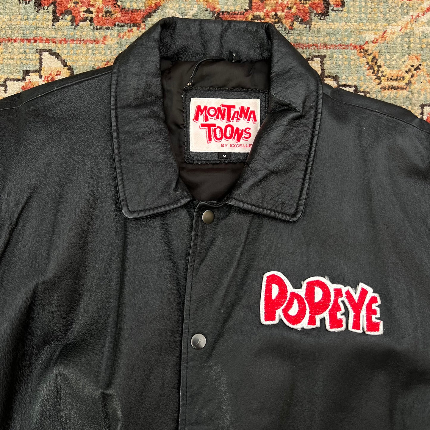 A black leather jacket with a pointed collar, snap-button front, and red embroidered 'Popeye' logo on the chest, laid flat on a patterned rug. The inner neck label reads 'Montana Toons by Cartoon'.