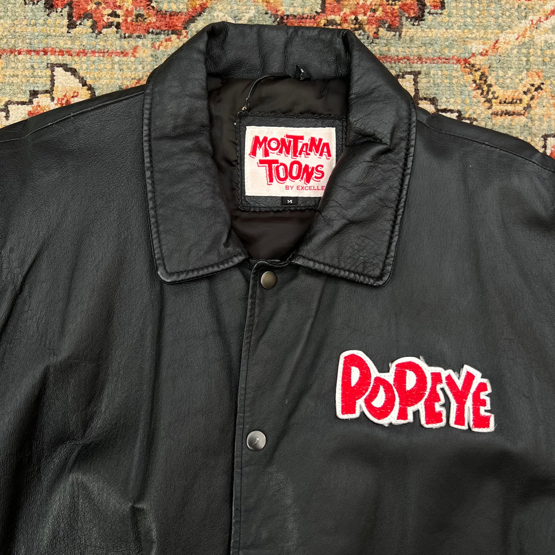 A black leather jacket with a pointed collar, snap-button front, and red embroidered 'Popeye' logo on the chest, laid flat on a patterned rug. The inner neck label reads 'Montana Toons by Cartoon'.