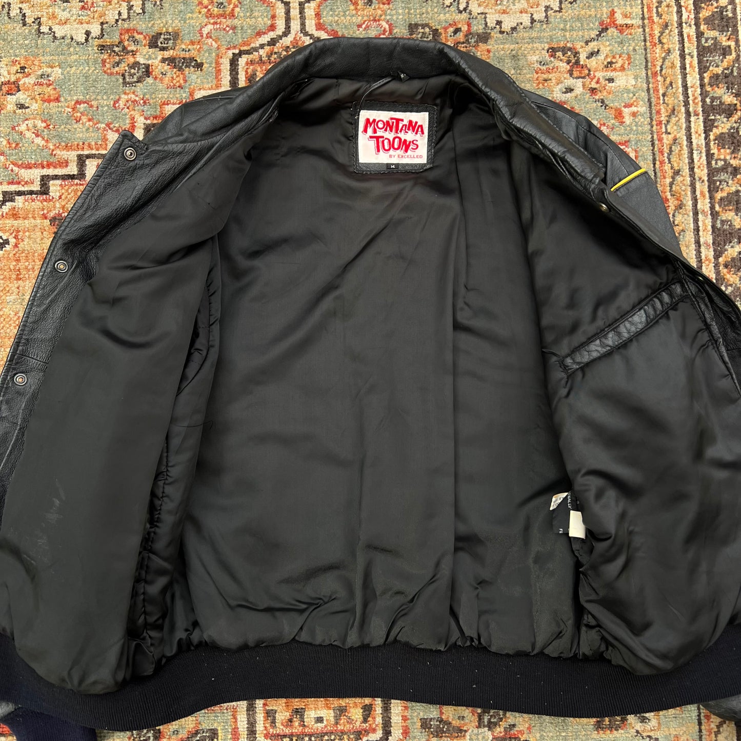 A black leather jacket with a white neck label reading 'Montana Today' in red, laid flat on a patterned rug.