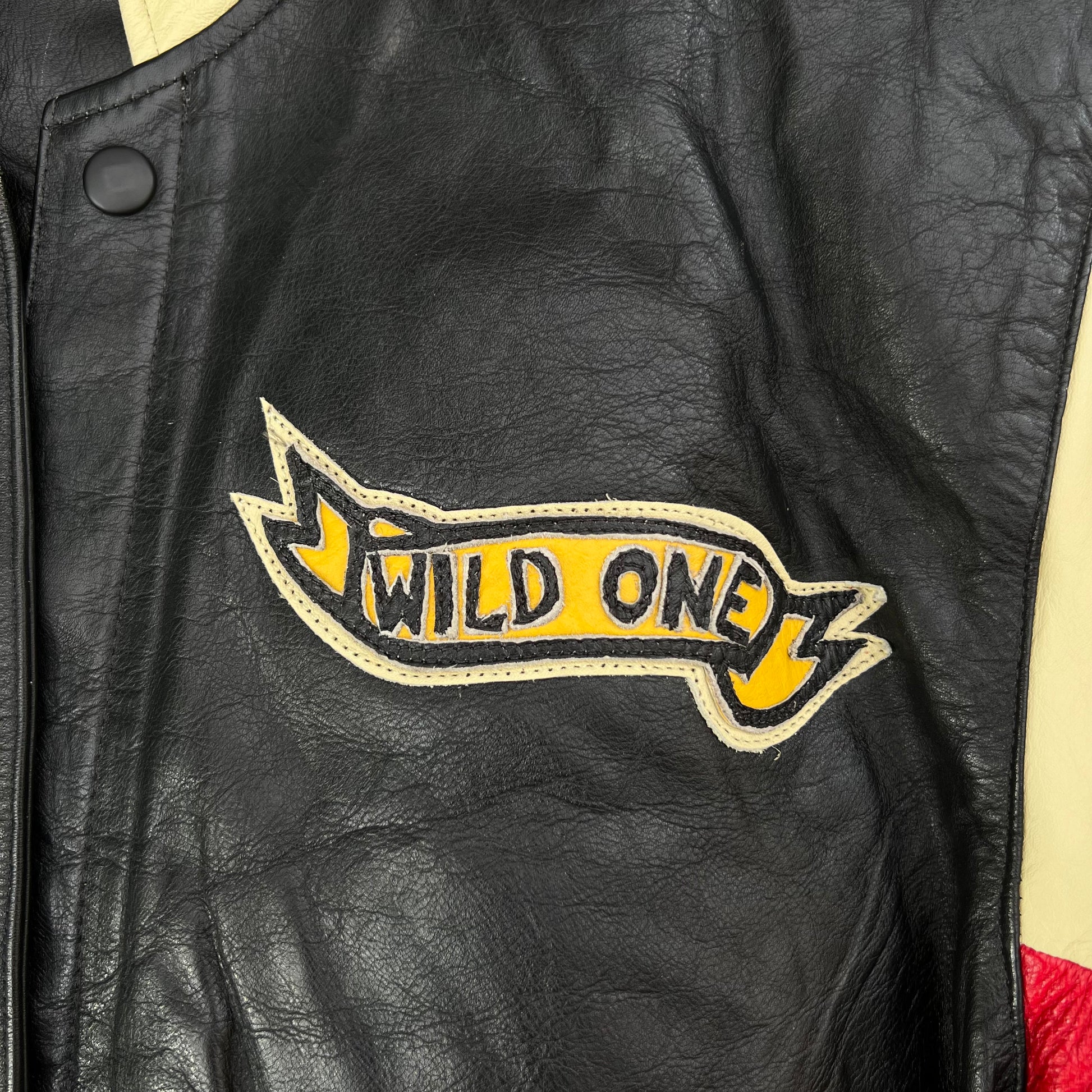 Close-up of a black leather jacket featuring a yellow and beige embroidered banner patch on the chest with the text 'WILD ONE'.