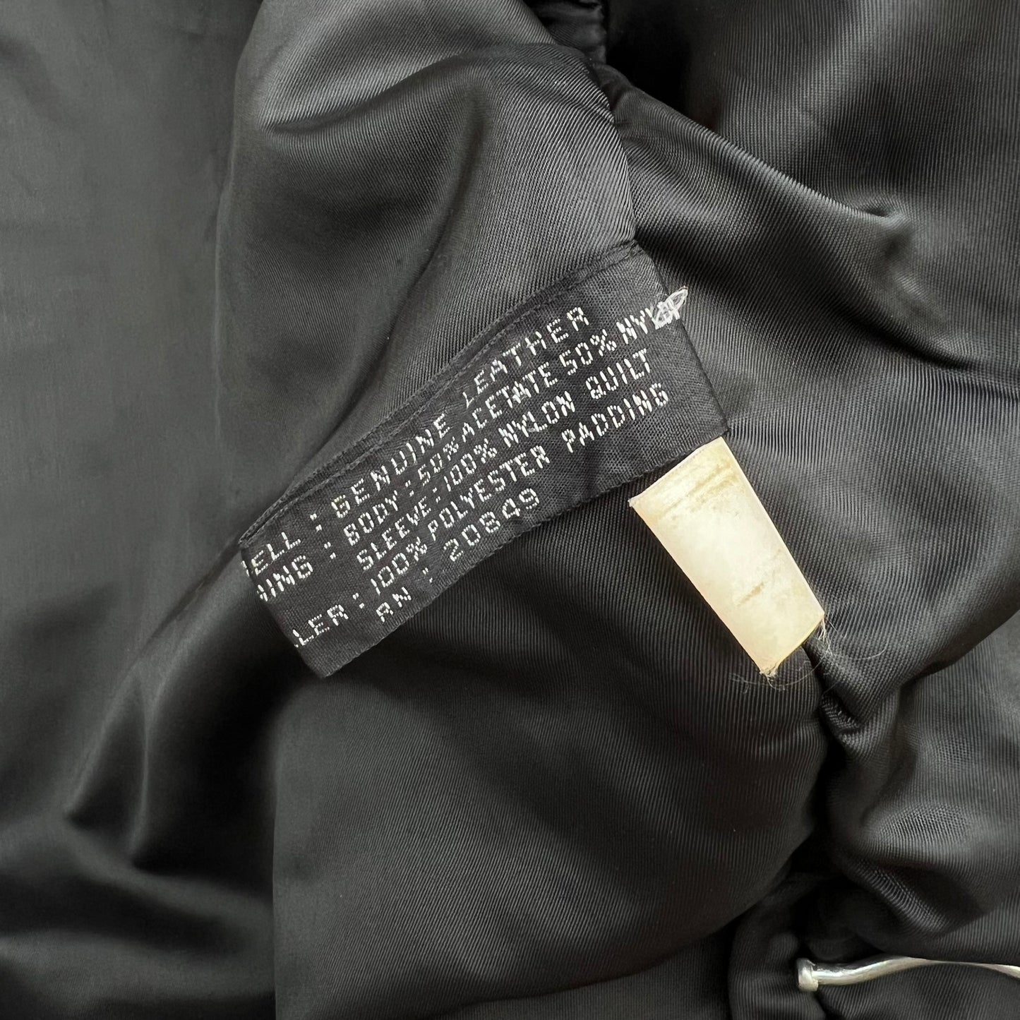 Close-up of a black padded garment interior showing a sewn-in label that reads 'Genuine Leather' and '100% Polyester Padded,' with a small beige hangtag attached.
