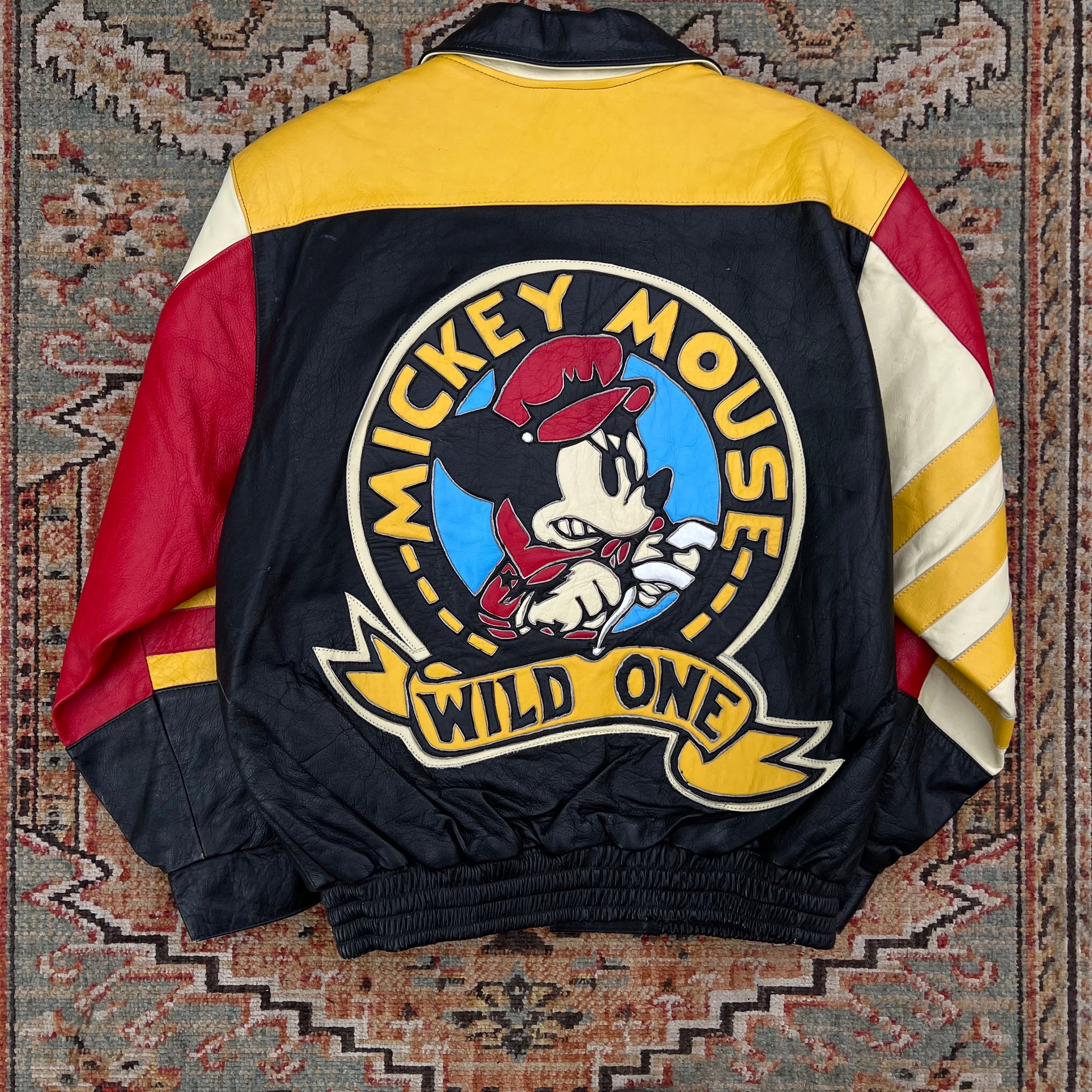 A vintage Mickey Mouse 'Wild One' colorblock jacket laid flat on a patterned rug. The back of the jacket features a large circular Mickey Mouse graphic with the text 'MICKEY MOUSE WILD ONE' in yellow and white.